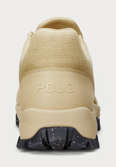 Beige színű sportcipő, amely texturált szövet felsőrésszel, gumitalppal rendelkezik, fekete pöttyökkel, és a saroknál feltűnő "POLO" logó található.