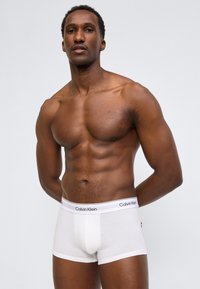 Μυώδης άνδρας που στέκεται με τα χέρια πίσω από την πλάτη, φορώντας λευκά εσώρουχα boxer Calvin Klein, μπροστά σε απλό ανοιχτό γκρι φόντο.