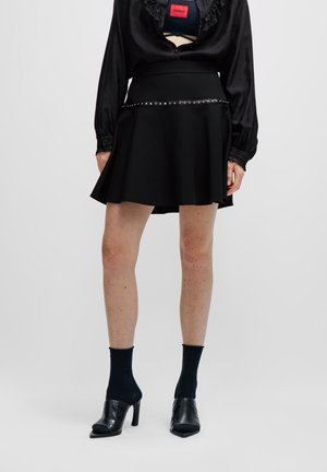 Femme portant une jupe noire évasée avec ceinture cloutée, une blouse noire à manches longues, des chaussettes noires aux chevilles et des chaussures noires à talons.