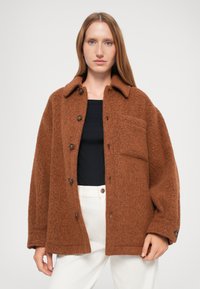 Giacca in misto lana marrone con finitura tessurizzata, colletto ampio, bottoni frontali e taschino sul petto, abbinata a un top nero e pantaloni bianchi.