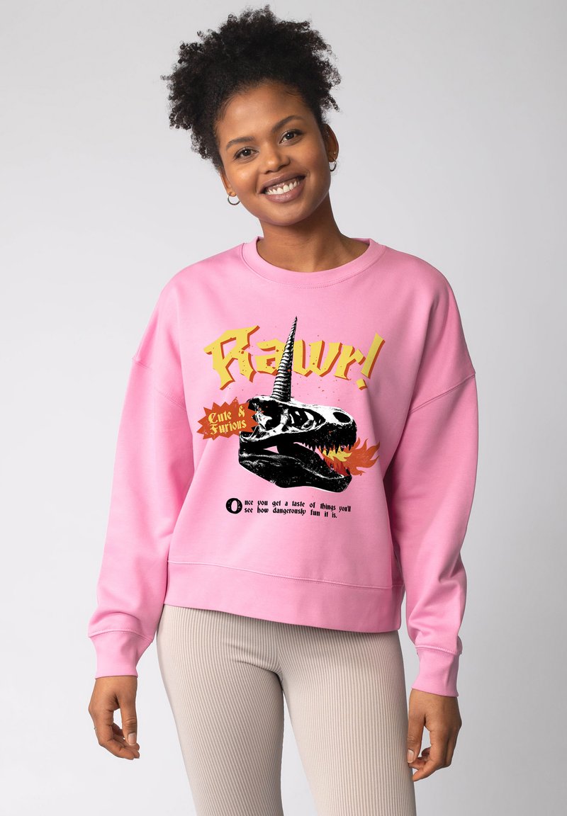 Roze katoenen sweatshirt met een zwarte dinosaurus schedel met een eenhoorn hoorn grafiek, gele en oranje tekst. Zachte textuur, losse pasvorm.