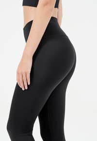 Zwarte high-waisted leggings van rekbaar materiaal, met een gladde textuur en een aansluitend ontwerp, zijwaarts afgebeeld met een hand op de heup.