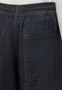 Pantalon en jean bleu foncé avec ceinture élastique et grandes poches arrière mises en valeur par des coutures orange contrastées.