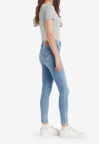 Kvinna klädd i en ljusgrå kortärmad topp, ljusblå tajta jeans, vita strumpor och vita sneakers, stående i sidoprofil.