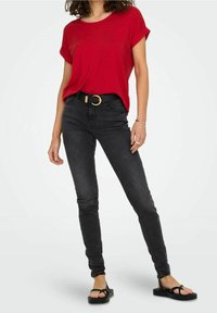 Kvinde iført en rød kortærmet top, sorte tætsiddende jeans med et sort bælte og sorte sandaler, stående mod en hvid baggrund.