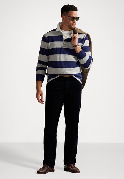 Marineblauw en grijs gestreepte rugbyshirt met een witte kraag, donkere broeken en bruine schoenen; houdt een beige jas vast; draagt een zonnebril.