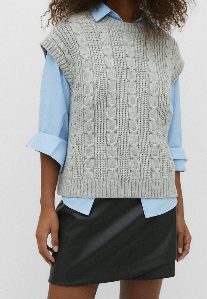 Femme portant un pull sans manches gris en maille torsadée par-dessus une chemise bleu clair retroussée et une jupe en cuir noire, debout devant un fond blanc.