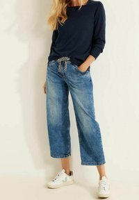 Losse lichtblauwe spijkerbroek met een geruite trekkoord, gecombineerd met een marineblauwe top met lange mouwen en witte sneakers.