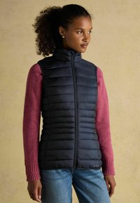 Melnā pūkainā veste ar vertikālām šuvēm, augsto apkakli un rāvējslēdzēja aizdari. Valkāta virs rozā džempera ar teksturētām piedurknēm. Ikdienas džinsu bikses.