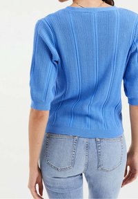 Modrý žebrovaný svetr s krátkými rukávy, s texturovaným vzorem, přiléhavého střihu a kulatým výstřihem, nošený s světle modrými džínami.