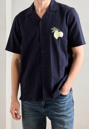Homme portant une chemise bleu marine à manches courtes avec boutons, en tissu texturé et une broderie jaune de citron sur la poitrine gauche, associée à un jean bleu.