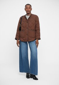 Giacca trapuntata marrone con colletto rotondo, due grandi tasche frontali e dettagli neri, abbinata a jeans in denim blu a gamba larga e stivali neri.