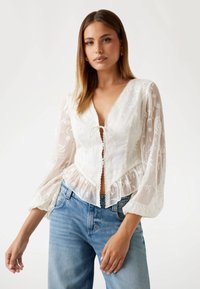 Witte geborduurde blouse met doorschijnende puffmouwen, een V-halslijn en een gerimpelde zoom; gecombineerd met lichtblauwe spijkerbroeken.