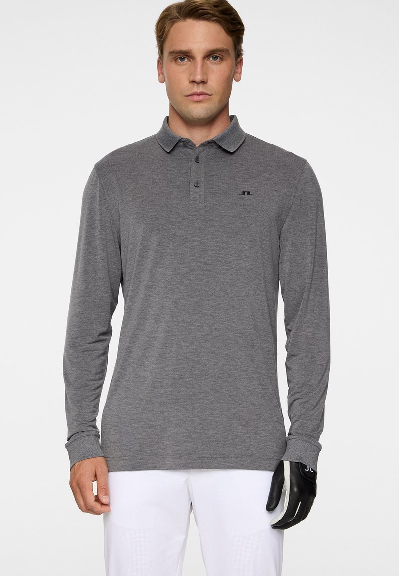 Mężczyzna ubrany w szarą koszulkę polo z długim rękawem i białe spodnie, na prawej ręce ma czarno-białą rękawiczkę, stoi przodem.