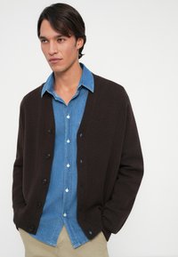 Mörkbrun cardigan med en avslappnad passform, som har sex svarta knappar, lager-på-lager över en ljusblå denimskjorta. Mjuk textur med vida ärmar.