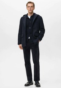 Joven vestido con un abrigo con capucha azul marino, un cárdigan abotonado, pantalones negros y zapatos de vestir negros, de pie contra un fondo blanco liso.