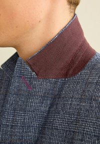 Maßgeschneiderte Jacke aus strukturiertem, blauem Stoff mit Karomuster, ergänzt durch einen bordeauxroten Kragen und lila Steppnähten.