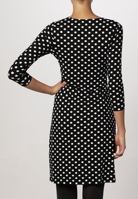 Svart klänning med vita prickar, långa ärmar, åtsittande midja, knälång fåll, mjukt tyg, slät textur, enkel rund hals.