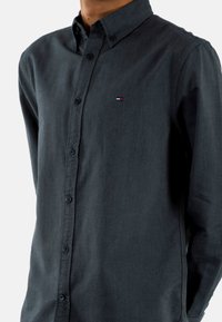 Camisa abotonada de tela gris oscuro, con cuello clásico, botones en la parte delantera y un pequeño logo rojo, blanco y azul en el pecho.