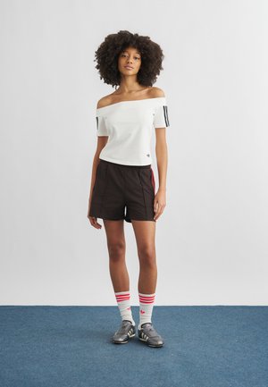 Kihara juustega naine, kes kannab valget õlgadeta topi, musti pükse punaste triipudega, valgeid sokke punaste triipudega ja musti tossusid, seistes.