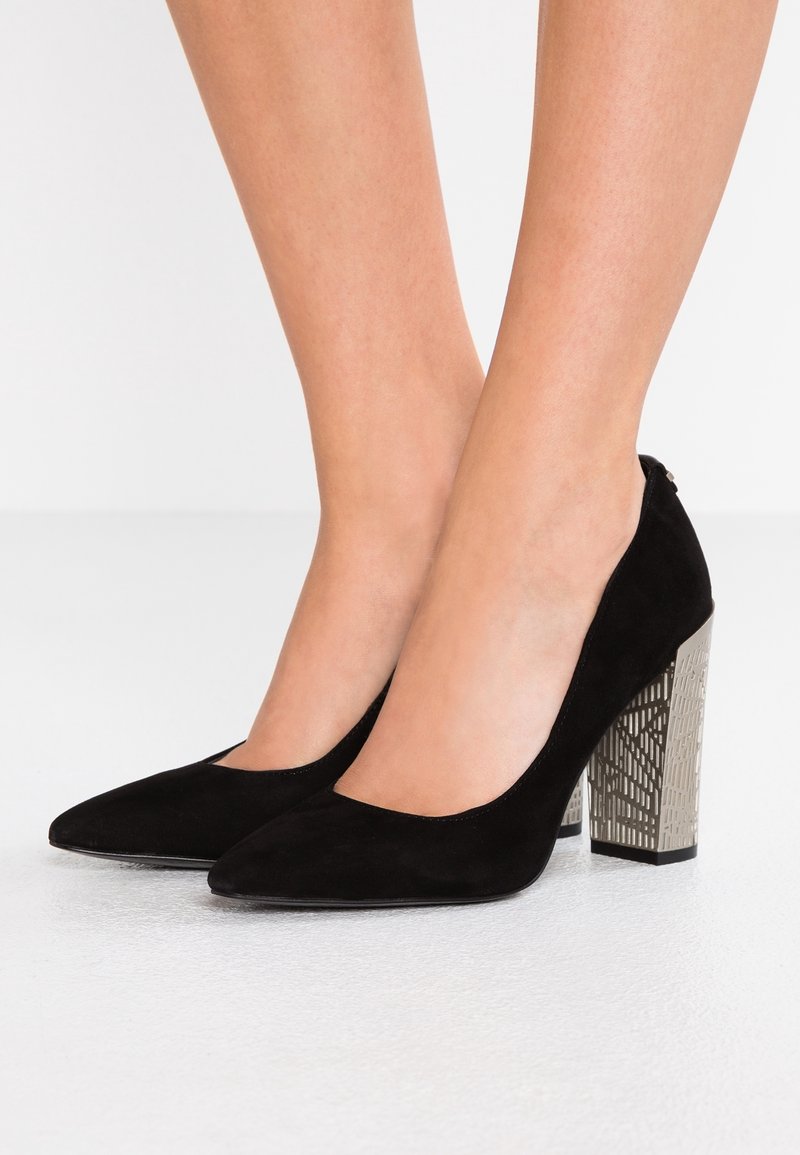 Escarpins en daim noir avec un bout pointu et un talon bloc métallique décoratif à motifs. Texture lisse et design élégant.