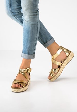 Fødder iført gyldne metalliske platformsandaler med ankelremme og spænder, kombineret med opsmøgede lyseblå jeans på hvid baggrund.