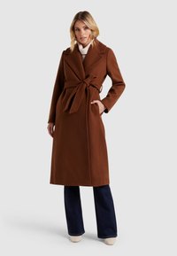 Brun, mellemlang frakke i et blødt materiale med bælte i taljen, store revers og sidelommer. Båret over flagrende jeans og lyse sko.