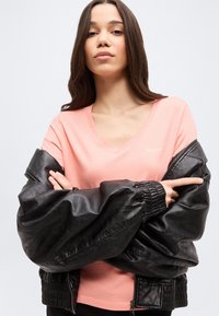 Νέα γυναίκα με μακριά σκούρα μαλλιά, σταυρωμένα τα χέρια, ντυμένη με ροζ μπλούζα με V-λαιμόκοψη και μαύρο δερμάτινο μπουφάν περασμένο στους ώμους.