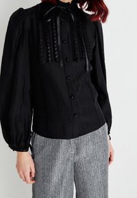 Blouse noire à manches longues avec des rayures verticales, détail froncé sur le devant et nœud en satin au cou. Associée à un pantalon rayé gris.