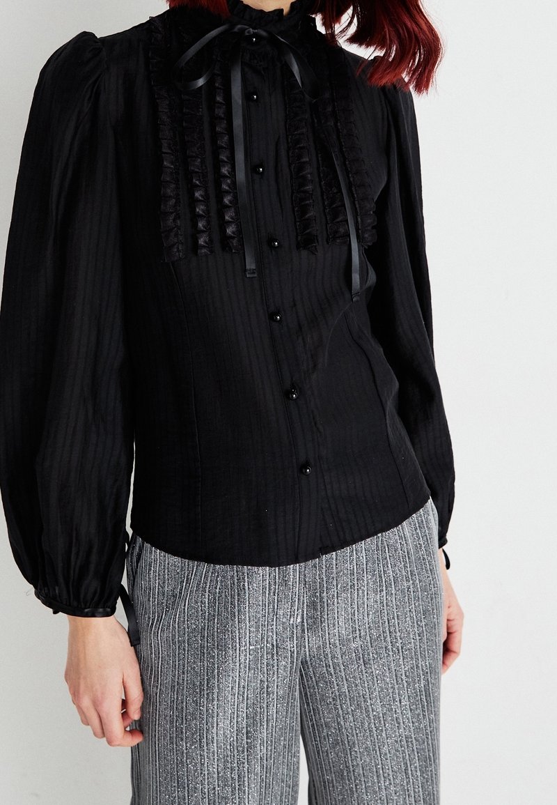 Blouse noire à manches longues avec des rayures verticales, détail froncé sur le devant et nœud en satin au cou. Associée à un pantalon rayé gris.