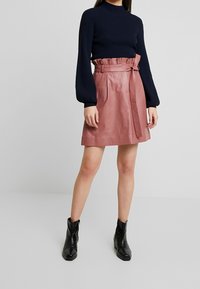 Vaaleanpunainen nahkahame, jossa on korkea vyötärö, röyhelövyö ja sivutaskut, yhdistettynä tummansiniseen ribbiknitti-paitaan ja mustiin nilkkureihin.