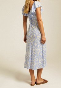 Sininen kukallinen midi-mekko, jossa on röyhelöhihoja ja solmittu selkäosa; mekko sisältää pieniä keltaisia kukkia vaaleansinistä taustaa vasten.