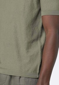 Top corto a maniche corte in tessuto terry verde con orlo a coste e spacchi laterali. Superficie textured e vestibilità rilassata, con braccia e spalle visibili.