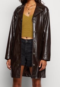 Žena v tmavě hnědém kožichu s kroko vzorem přes hořčicový pletený crop top a černé džíny s vysokým pasem, doplněná o vrstvené zlaté náhrdelníky.