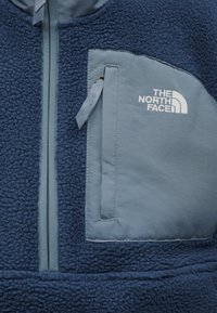Μπλε φλις μπουφάν με φερμουάρ στην τσέπη του στήθους από πιο ανοιχτό μπλε ύφασμα, με το λευκό λογότυπο "THE NORTH FACE".