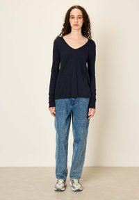 Moteris su garbanotais plaukais dėvi juodą ilgomis rankovėmis V formos kaklo marškinėlį, laisvus mėlynus džinsus ir baltus sportinius batelius, stovėdama priešais paprastą smėlio spalvos sieną.