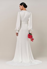 Langärmliges weißes Kleid mit glatter Textur, taillierter Silhouette und verlaufendem Saum. Ausgestattet mit einem passenden Gürtel und einem Blumenstrauß in der Hand.