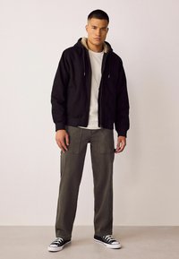 Veste aviateur noire à capuche avec une doublure en polaire beige, associée à un pantalon cargo vert olive et des baskets noires avec des lacets blancs.