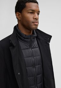 Uomo che indossa una giacca nera trapuntata con zip, stratificata sotto un cappotto di lana nero con il colletto alzato, su uno sfondo chiaro e semplice.