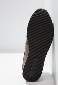 Scarpa in suede beige con suola in gomma testurizzata, caratterizzata da scanalature orizzontali e un dettaglio di branding. La scarpa è posizionata in verticale contro un muro.