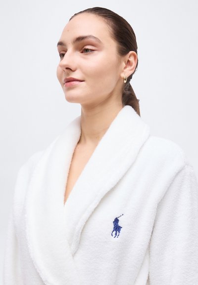 Γυναίκα με καστανά μαλλιά δεμένα πίσω, φορώντας λευκή ρόμπα μπάνιου με μπλε κεντημένο λογότυπο αλόγου, στραμμένη ελαφρώς προς τα αριστερά σε απλό φόντο.