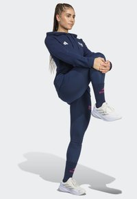 Kvinde i marineblå sportstøj og hvide sneakers, der strækker sig ved at kramme det ene knæ til brystet, stående på ét ben mod en ensfarvet baggrund.
