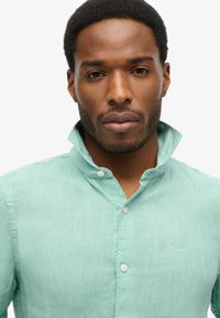 Miętowa lniana koszula z kołnierzykiem zapinanym na guziki, z delikatnym logo na piersi i guzikiem z przodu.
