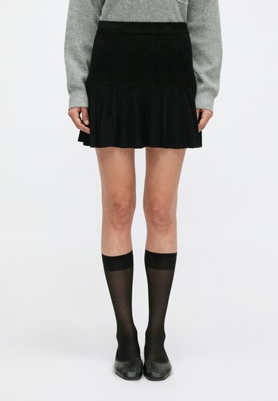 Schwarzer, plissierter Rock aus einem weichen Material, mit einem figurbetonten Bund. Getragen mit schwarzen, knielangen Socken und schwarzen Ballerinas, vor einem weißen Hintergrund.
