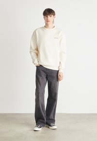 Sudadera de color crema claro con escote redondo y un pequeño logo, combinada con unos jeans de pierna ancha en gris oscuro y zapatillas negras. Texturas de tela suaves.