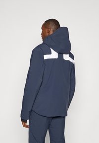 Casaco impermeável azul-marinho com capuz, apresentando detalhes brancos nos ombros e uma textura exterior suave. Bolsos com fecho éclair nas laterais.