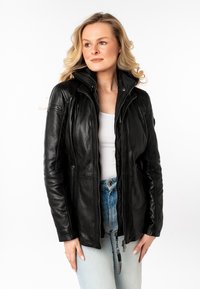Vrouw met blond haar in een zwarte leren jas over een witte top en lichtblauwe jeans, die enigszins opzij kijkt tegen een witte achtergrond.