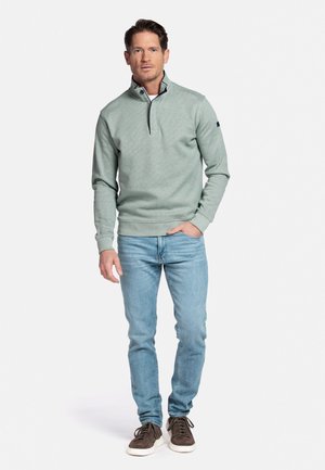 Mann trägt einen hellgrünen Pullover mit Viertelreißverschluss, hellblaue Jeans und braune Turnschuhe und steht mit einer Hand in der Tasche.