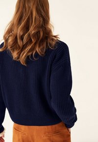 Kobieta o falowanych, rudych włosach, ubrana w granatowy sweter z dzianiny i brązowe spodnie, pokazana od tyłu na beżowym tle.