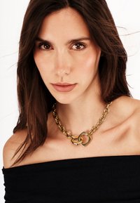 Guldkedjehalsband med länkdesign och två sammanlänkade geometriska former. Bärs på en bar axel, vilket framhäver dess textur.